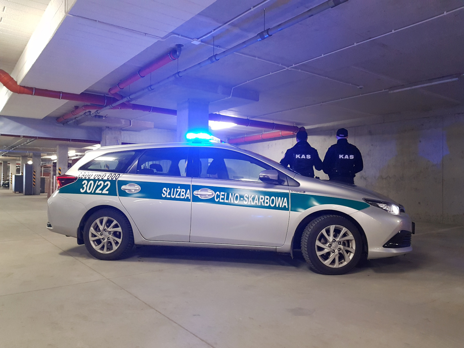 na zdjęciu dwóch funkcjonariuszy celno-skarbowych odwróceni plecami do zdjęcia, za nimi samochód służbowy z opisem służba celno-skarbowa