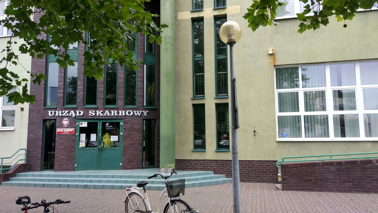 Na zdjęciu budynek w szaro - zielonej elewacji, wejście główne elewacji wyłożone płytkami w kolorze brązowawym, obramowanie okien i drzwi w kolorze ciemno zielonym