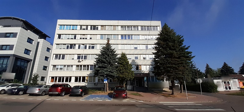 Na zdjęciu wielokondygnacyjny budynek w kształcie prostokąta będący siedziba Delegatury. Przed budynkiem parking z wyznaczonym miejscem postojowym dla osoby niepełnosprawnej.