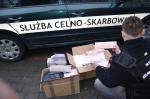 Funkcjonariusz Służby Celno-Skarbowej wyjmuje z pudeł kartony papierosów bez polskich znaków akcyzy. Pudła leżą obok radiowozu Służby Celno-Skarbowej.