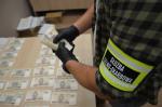 Pracownik KAS trzyma w dłoniach banknoty, więcej banknotów leży na stole przed nim.