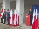 Uroczystość z udziałem mundurowych oraz mężczyzny przemawiającego przy mównicy z logo Krajowej Administracji Skarbowej, otoczona polskimi flagami.