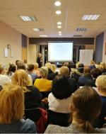 Grupa przedsiębiorców siedząca na sali konferencyjne. W tle osoba prowadząca szkolenie